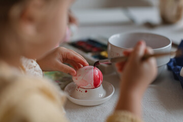 Close-up of a little girl painting eggs for Easter at a table in a cozy kitchen. Easter family traditions.