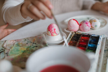 Close-up of a young mom painting eggs with a cute little daughter at a table in a cozy kitchen. Easter family traditions.