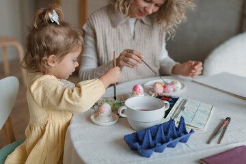 Obraz na płótnie Canvas A cute little girl painting eggs with her young stylish mom at a table in a cozy kitchen. Easter family traditions.