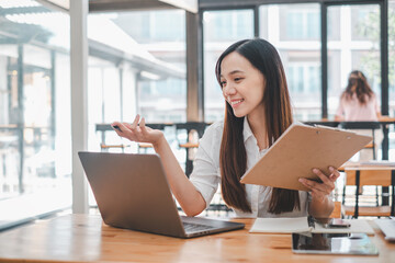 Business analytics concept, Smiling Asian businesswoman multitasking with a laptop and clipboard in a well-lit cafe, indicating a discussion or presentation.