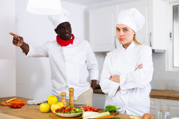 Dissatisfied adult chef controlling work of young female assistant in kitchen