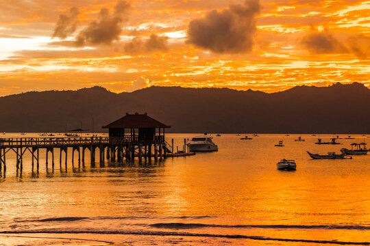 A wooden pier stretches out into a calm bay at sunset, with mountains in the background.