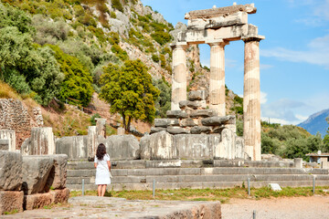 A long hair woman in a white dress looking at the ancient temple complex of Athena Pronaia in Delphi. Sunny day, blue sky. The archaeological site, UNESCO World Heritage Site, Delphi, Greece.
