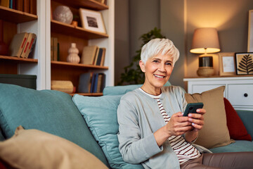 A portrait of a smiling senior woman using her smart phone and relaxing on a couch in the living...