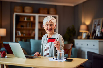 Fototapeta na wymiar A portrait of a happy senior adult woman holding a credit card and sitting at a desk in front of a laptop ready to make an investment online