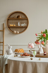 Interior design of easter dining room interior with round table, shelf on wall, colorful eggs, easter cake, hare sculptures, vase with tulips, lychee and personal accessories. Home decor. Template.