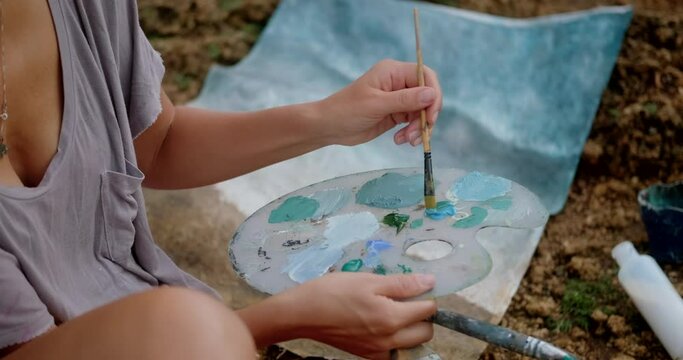 Close-up of a woman's hands mixing blue and green shades of acrylic paint with a brush on a palette and painting a picture of the ocean on canvas. The artist paints a picture outdoors.