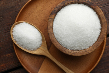 Granulated sugar in bowl and spoon on wooden table, top view