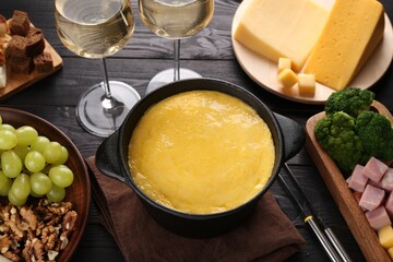 Fondue pot with melted cheese, glasses of wine and different products on black wooden table, above view