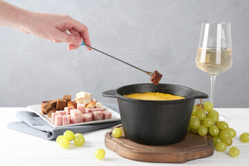 Woman dipping piece of bread into fondue pot with tasty melted cheese at white wooden table against...