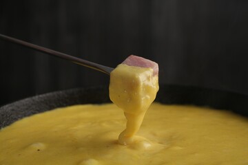 Dipping piece of ham into fondue pot with tasty melted cheese against dark gray background, closeup