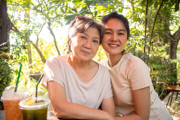Asian Thai Chinese elder mother and daughter cuddling and hug, looking at camera, drinking beverages at cafe garden, spend time together.