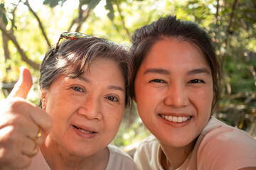 Asian Thai Chinese elder mother and daughter cuddling and hug, looking at camera, drinking beverages at cafe garden, spend time together.