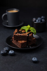 Chocolate brownie cake with mint, blueberries, chocolate topping and espresso. Traditional American dessert. Popular cake with dark chocolate on a black background. Close-up, selective focus.