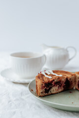 a round croissant on a green plate one cut in half.Nearby are a napkin with red patterns and cutlery,a cup of coffee,a milk jug. White background.