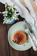 in a plate there is a round New York croissant in out of focus, next to a blue vase with snowdrops in focus and a striped towel with cutlery on a dark wooden background, shot from above