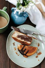 in a plate there is a round New York croissant with chocolate cut in half, next to it there is a blue vase with snowdrops, a cup of coffee and a striped towel with cutlery on a dark wooden background