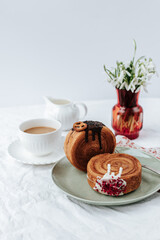 There is a two round croissant on a beige plate.Next to it are a napkin with red patterns and vintage cutlery,a cup of coffee, a milk jug and a red vase with snowdrops.White backgrounds
