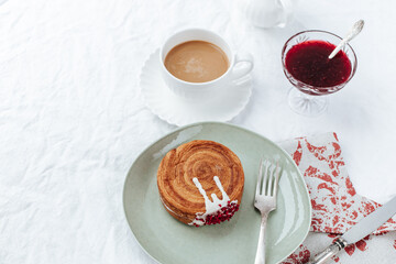 There is a round croissant on a green plate.next to it is a napkin with red patterns and vintage cutlery, a cup of coffee,a milk jug and a glass bowl of jam.White background