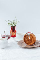 There is a round croissant on a beige plate.Next to it are a napkin with red patterns and vintage cutlery,a cup of coffee, a milk jug and a glass bowl of jam, red vase with snowdrops.White backgrounds