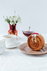 There is a round croissant on a beige plate.Next to it are a napkin with red patterns and vintage cutlery,a cup of coffee, a milk jug and a glass bowl of jam, red vase with snowdrops.White backgrounds