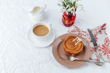 There is a round croissant on a beige plate.Next to it are a napkin with red patterns and vintage cutlery,a cup of coffee, a milk jug and a glass bowl of jam, red vase with snowdrops.White backgrounds