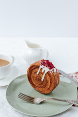 There is a round croissant on a green plate.next to it is a napkin with red patterns and vintage cutlery, a cup of coffee,a milk jug .White background