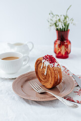 There is a round croissant on a beige plate.Next to it are a napkin with red patterns and vintage cutlery,a cup of coffee, a milk jug and a red vase with snowdrops.White backgrounds