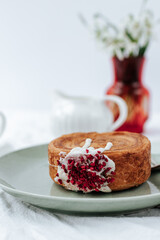 There is a round croissant on a green plate.next to it is a napkin with red patterns and vintage cutlery, a milk jug and a red vase with snowdrops.White background