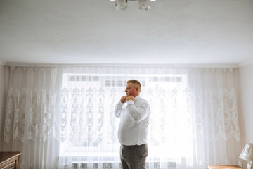 A man in a white shirt stands by the window in the room and fastens the buttons on his collar and sleeves. Watch on hand. Stylish business portrait of a man, close-up photo. The groom is preparing.