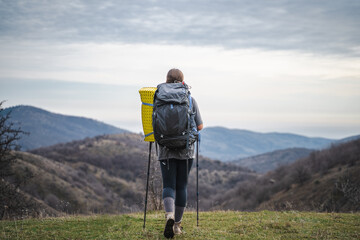 Back view of young backpacker woman with backpack on travel and enjoying mountains landscape, tourist in hiking equipment looks into distance at peaks of mount on a cloudy day, summer vacation journey