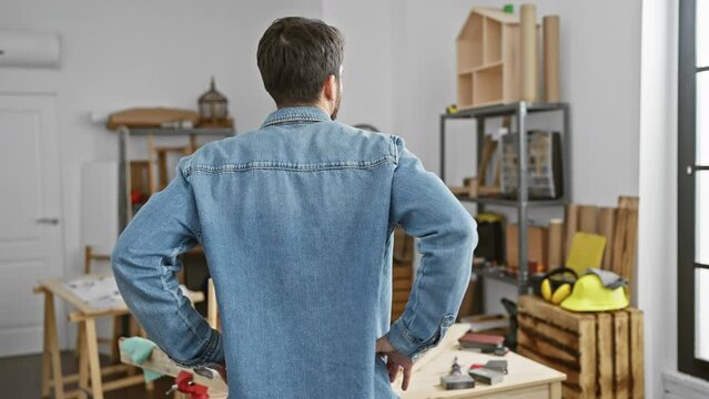 Rear view of a contemplative man wearing a denim jacket standing in a carpentry studio with workbench and tools.