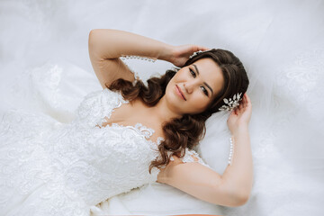 A brunette bride is lying on a lace dress and tiara, posing touching her hair