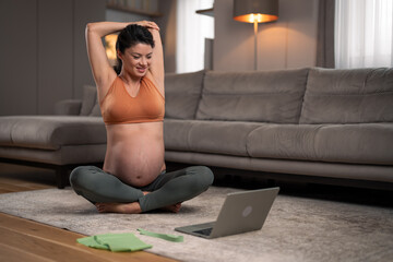 A beautiful and healthy pregnant woman stretching her arms an watching a video tutorial on a laptop at home