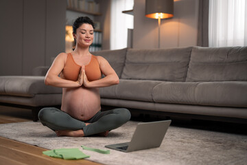 A satisfied young pregnant lady meditating at home while sitting on the floor in front of a laptop