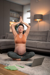A focused young pregnant woman meditating with clasped hands above her head sitting in the living room.