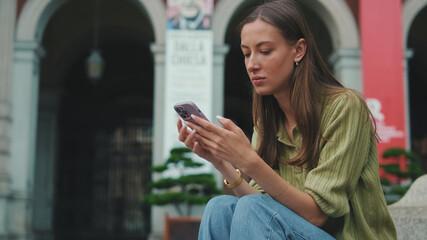 Young woman with brown hair, dressed in an olive green sweater, uses a phone while sitting on the square of the old city