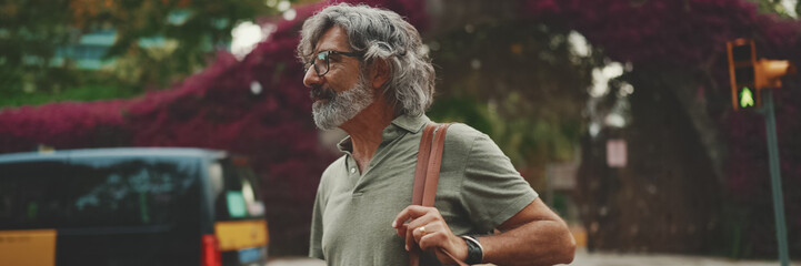 Friendly middle-aged man with gray hair and beard wearing casual clothes crosses the road at...