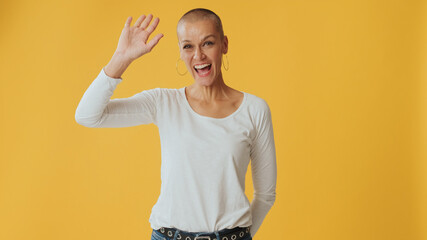Young hairless woman waving palm in greeting gesture looking at camera, isolated on yellow...