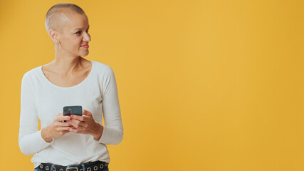 Smiling young hairless woman,with phone in hands, looking away at advertising space isolated on...