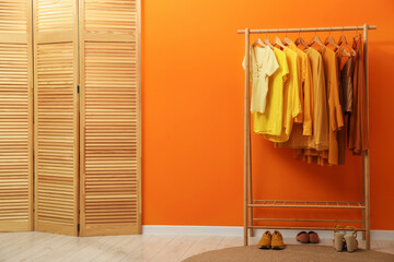Rack with different stylish women's clothes, shoes and wooden folding screen near orange wall indoors