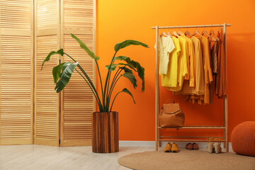 Rack with different stylish women's clothes, shoes, backpack and green houseplant near orange wall...