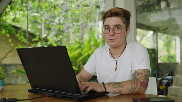 Transgender person with tattoos smiles, works on laptop in modern open-plan workspace. Joyful moment, gender inclusivity at job, relaxed office environment, tech usage. Slow motion.