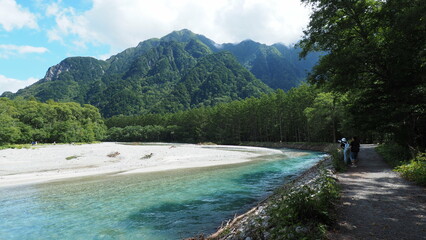 Azusa River with turquoise color water, withtravllers on trees covered trail by the right, surrounded by Japanese Alps mountains under a beautiful blue sky with white clouds