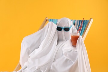 Person in ghost costume and sunglasses with glass of drink relaxing on deckchair against yellow...