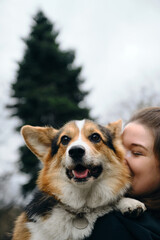 A young Caucasian woman holds and kisses a dog in arms in the park while walking. Female owner and a Welsh corgi pembroke tricolor. Life style portrait with sweet pet.