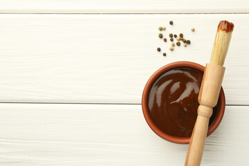 Tasty barbeque sauce in bowl, brush and peppercorns on white wooden table, top view. Space for text