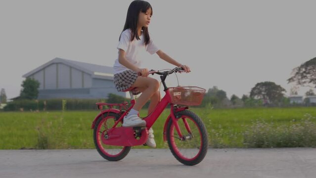 Video taken using a handheld camera Portrait Asian Thai kid girl, aged 8 to 10 years old, riding a red bicycle, playing on the road outdoors, happy and having fun. The background is a green field.