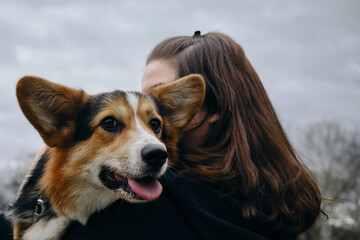 A young woman with brown hair is holding her cute corgi dog outdoors. The dog has its tongue out and looks happy to be hugged.