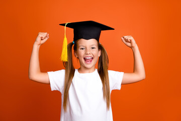 Photo of delighted positive schoolchild raise fists success wear mortarboard hat isolated on orange color background